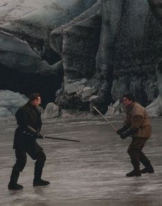 two men are playing with swords on the ice in front of some rocks and snow