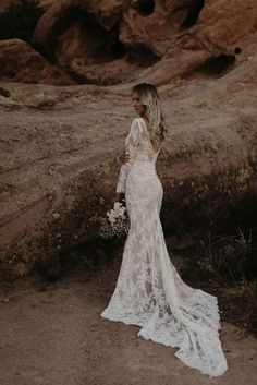 a woman standing in the desert wearing a white lace wedding dress and holding a bouquet