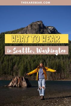 a woman in yellow jacket and blue jeans standing on the shore with her arms spread out