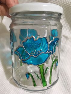 a hand holding a glass jar with blue flowers painted on the lid and bottom, sitting on a white surface