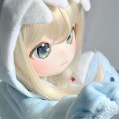 a doll with long blonde hair and blue eyes is wearing a white hooded jacket while holding a teddy bear