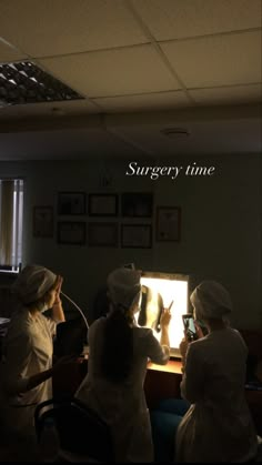 three women in white lab coats sitting at a table with an illuminated screen behind them that says surgery time