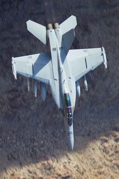 a fighter jet flying through the air over a dry grass covered field and dirt ground