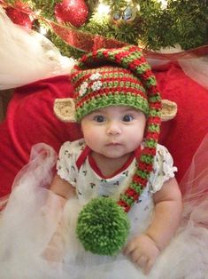 a baby wearing a knitted elf hat sitting in front of a christmas tree