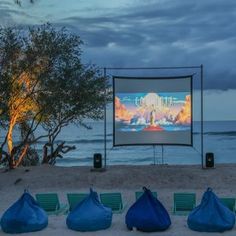 an outdoor movie is set up on the beach with chairs and blue bags around it