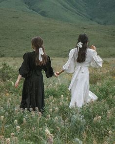 two girls in black and white dresses holding hands while walking through tall grass with mountains in the background