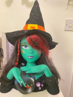 a doll with red hair and green skin wearing a witches hat on top of a counter