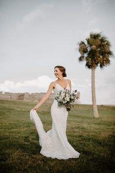 a woman in a wedding dress holding a bouquet and walking through the grass with palm trees behind her