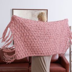 a woman holding up a pink crocheted shawl in front of a couch