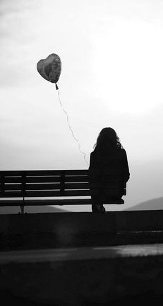 a person sitting on a bench with a heart shaped balloon in the sky above them