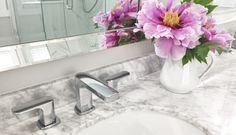 a white vase filled with purple flowers sitting on top of a bathroom counter next to a sink