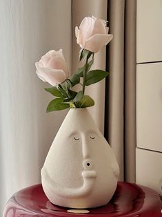 a white vase with two pink roses in it on top of a red table next to a window