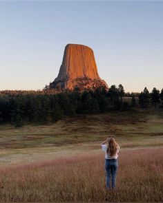 a woman standing in a field looking up at a tall mountain with trees on it