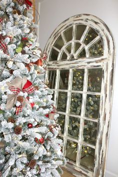 a white christmas tree with red and green bows in front of an arched window that is decorated with plaid ribbon