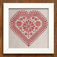 a cross stitch heart in a white frame on a wooden surface with a red border