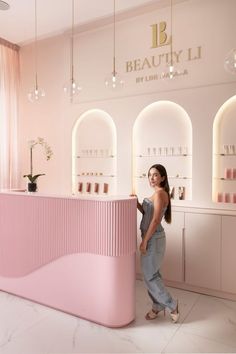 a woman standing next to a pink counter in a beauty shop with lights hanging from the ceiling