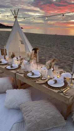 a table set up on the beach with candles and plates in front of an teepee tent