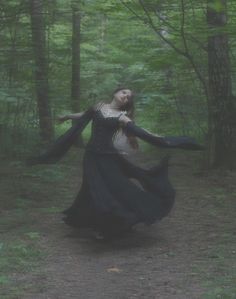 a woman is dancing in the woods with her arms spread out and she's wearing a long black dress