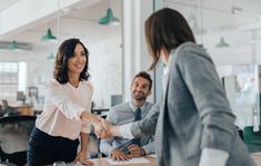 two business people shaking hands in an office with one woman sitting at a desk and the other standing up