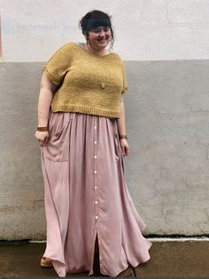 a woman standing in front of a white wall wearing a pink skirt and yellow sweater