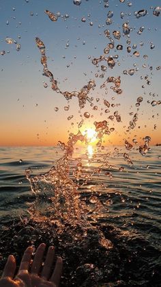 someone is holding their hand out to the water as the sun sets in the background