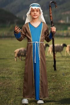 a young boy dressed up as a shepherd holding a stick and standing in the grass with sheep behind him