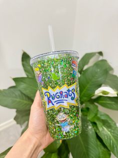 a person holding up a cup filled with green sprinkles next to a plant