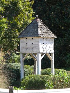 a wooden bird house sitting in the middle of a garden with hedges and trees around it