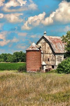 an old barn sits in the middle of a field with tall grass and trees around it