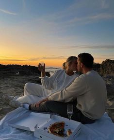 two people sitting on the beach eating pizza and drinking wine while watching the sun set