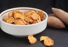 a white bowl filled with potato chips next to two wooden utensils on a black surface