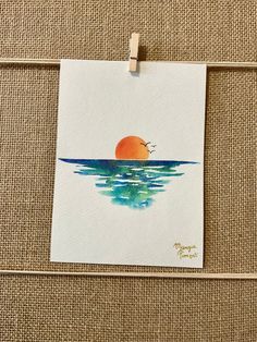 a piece of paper hanging on a clothes line with a painting of an orange sun in the water