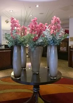 three silver vases with pink flowers in them on a table at a hotel lobby