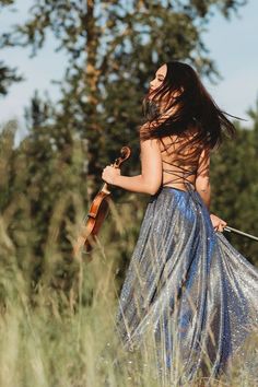 a woman in a blue dress is holding a violin and walking through tall grass with trees behind her