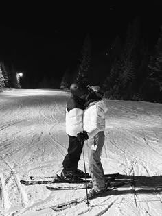 two people standing on skis in the snow at night time with their arms around each other