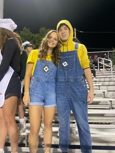 two people standing next to each other wearing overalls and yellow hoodies at a football game