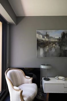 a white chair sitting in front of a window next to a painting on the wall