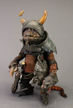 an action figure with horns and armor is posed on a gray surface, wearing a helmet