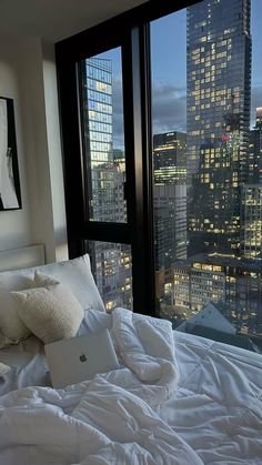 a bed with white sheets and pillows in front of large windows overlooking the city at night