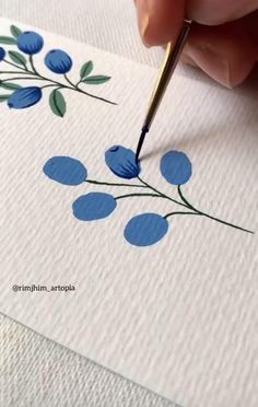 a person is using a pen to paint blue flowers on white paper with green leaves