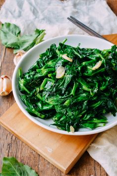 a white bowl filled with greens on top of a wooden cutting board next to garlic