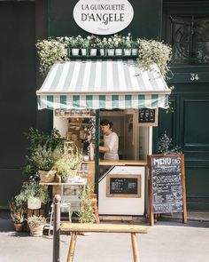 a man standing in front of a small store with plants on the outside and an awning over it