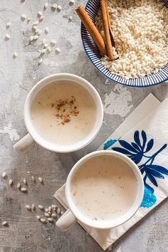 two mugs filled with coffee sit on a table next to some cinnamon sticks and rice