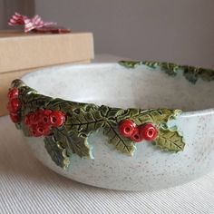 a white bowl with red berries and green leaves on it next to a wooden box