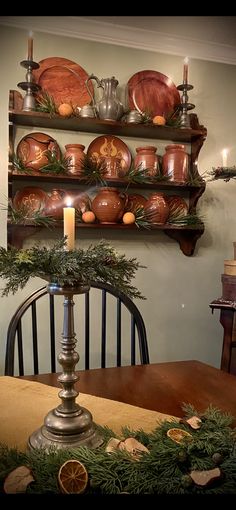 a dining room table with candles and plates on top of the shelves above it, surrounded by greenery