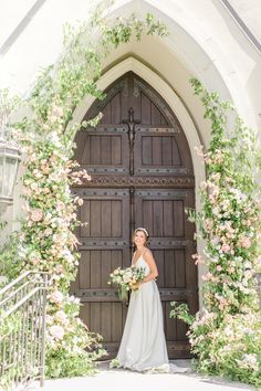 a bride standing in front of an arched doorway with flowers on it and holding her bouquet