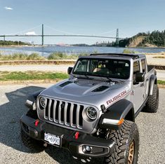 the jeep is parked on the side of the road by the water with a bridge in the background