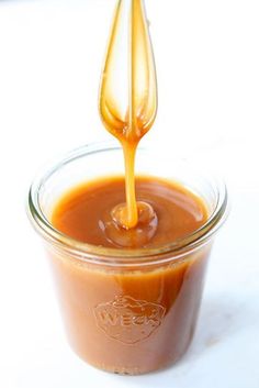 a spoon pouring sauce into a jar on a white surface with the lid open and an orange liquid in it