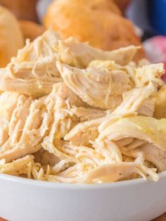 a white bowl filled with shredded chicken sitting on top of a wooden table next to bread rolls