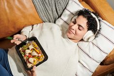 a woman laying on the couch with her headphones on and holding a plate of food